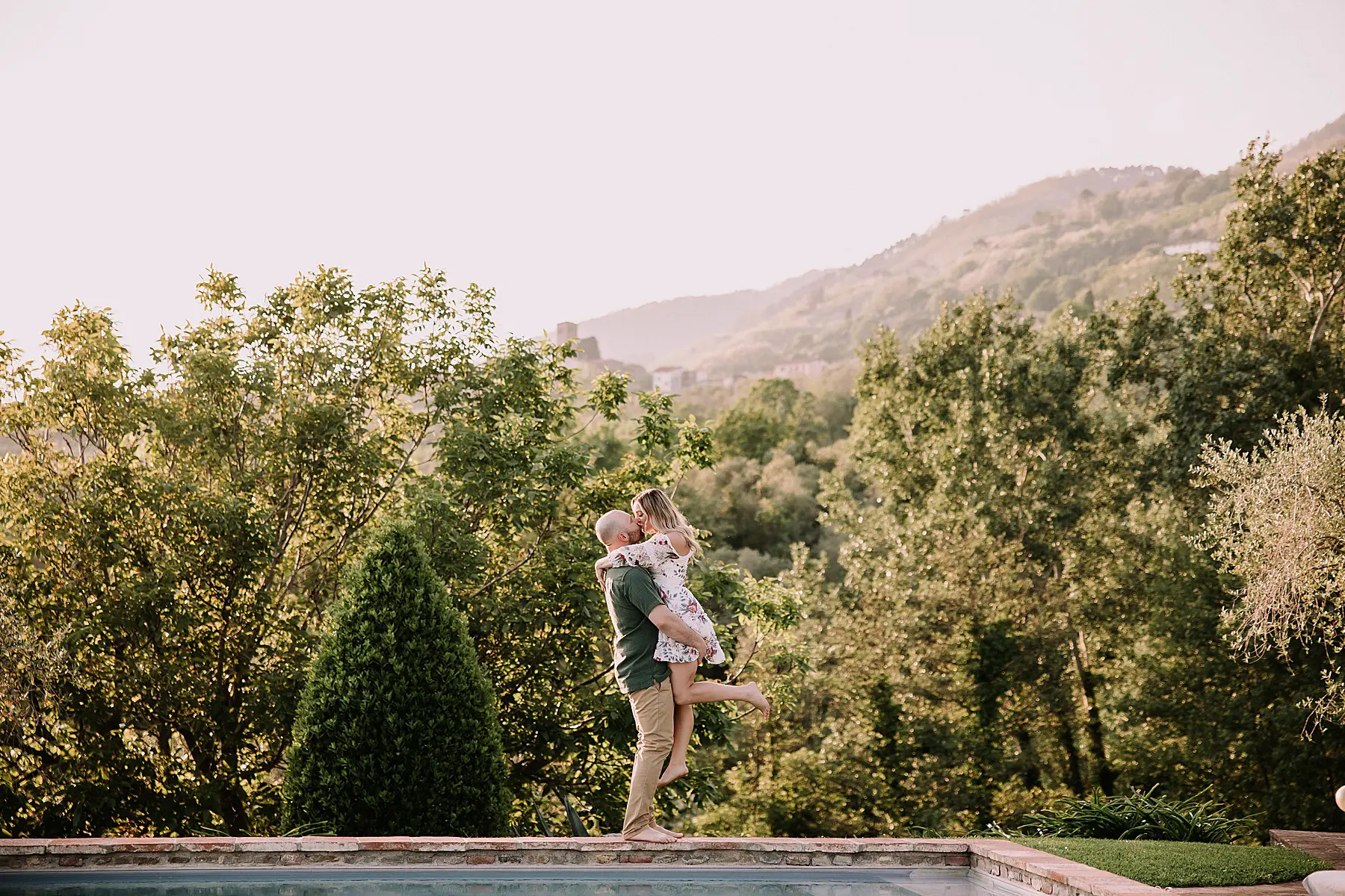 couple wearing formal attire standing next to a pool with the rolling hills of tuscany behind them. he is picking her up to give her a kiss