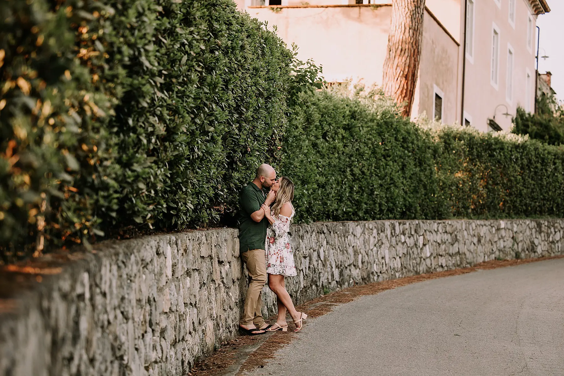 couple standing on an empty country road in tuscany. there is a rustic brick wall and a large native tree behind them. They are wearing formal attire and snuggling while posing for a photo