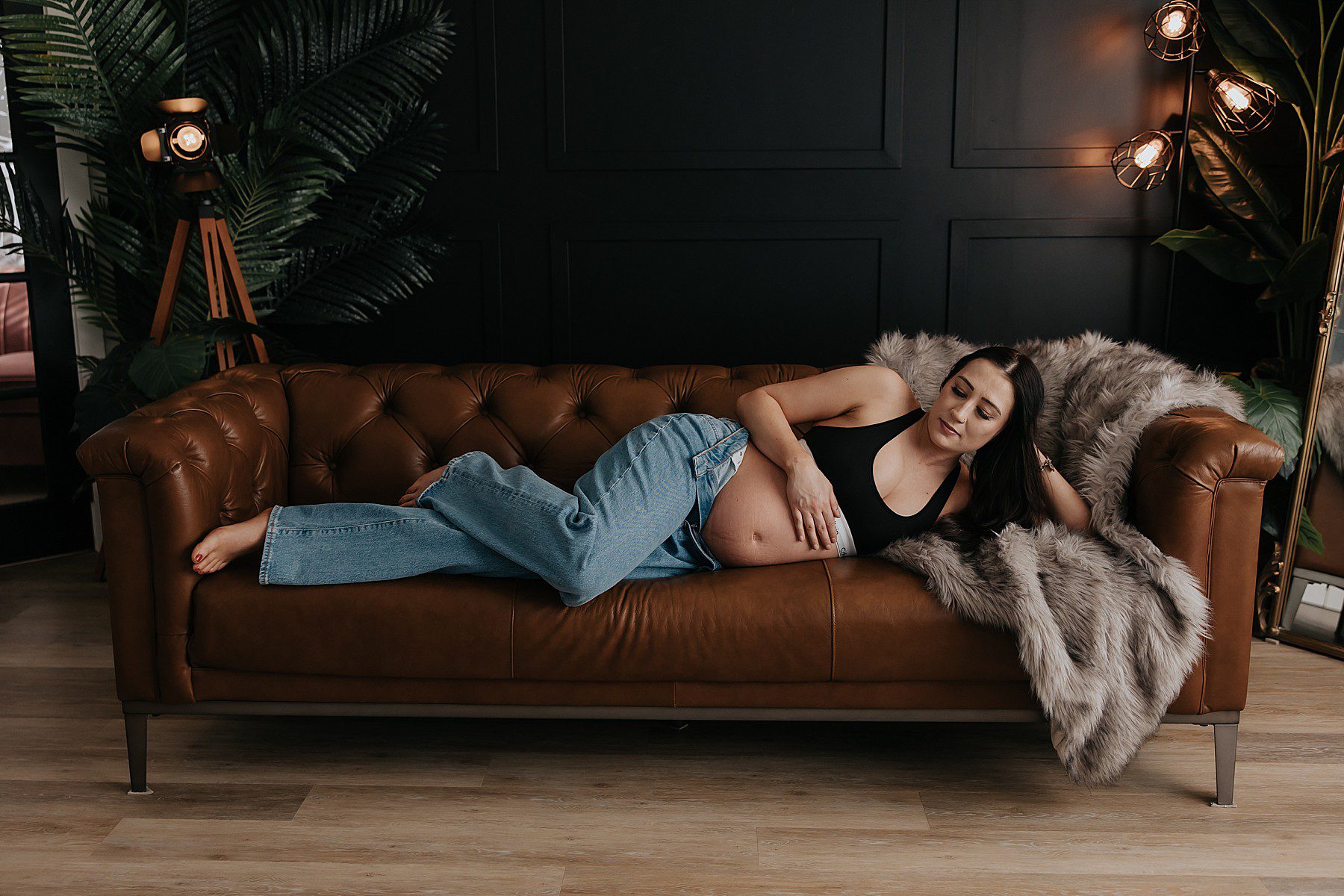 woman posing in a studio for maternity photos. She is wearing jeans with the top button undone and a black calvin klein bra