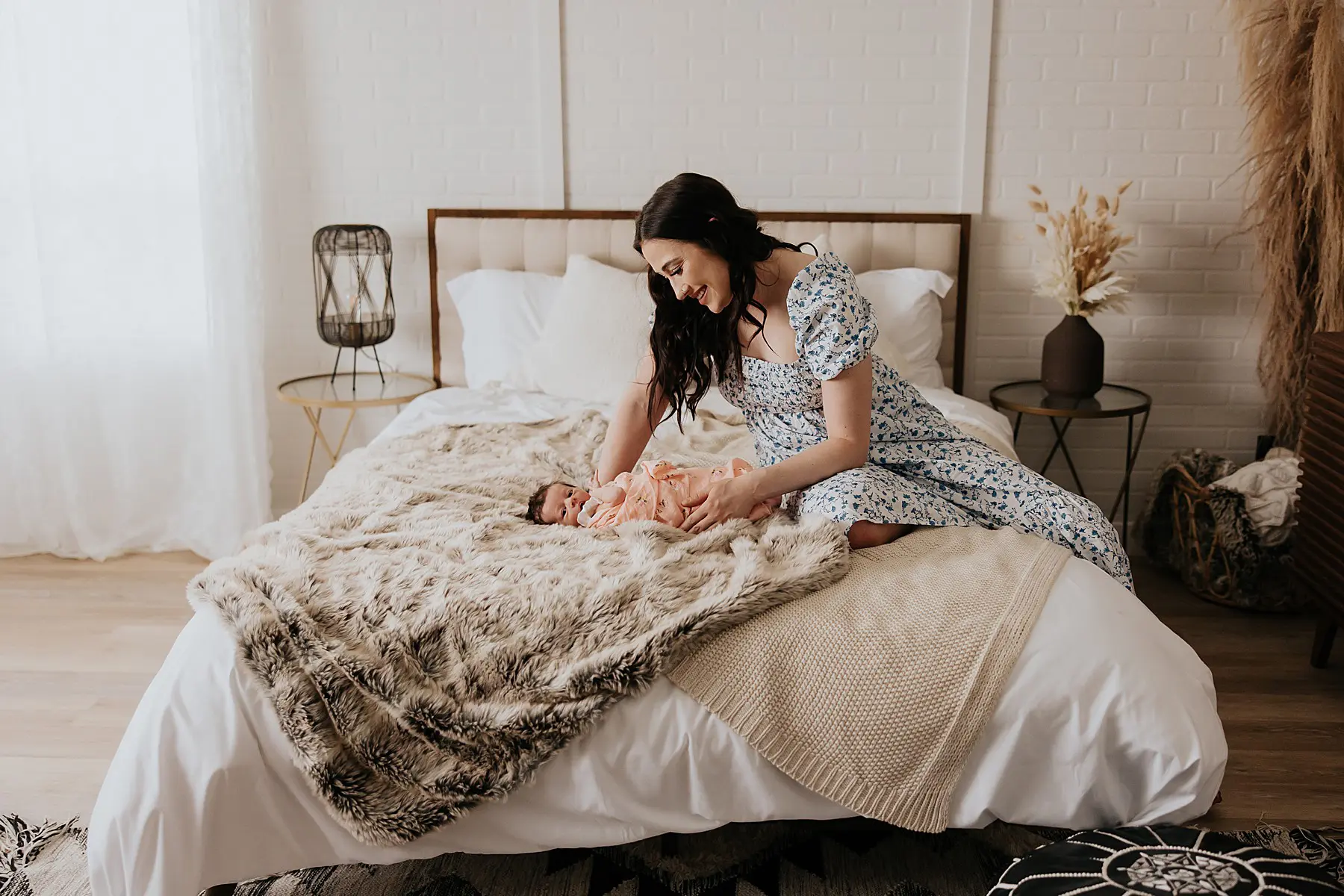 mom dressed in a white and blue dress. She is sitting on a bed and holding her newborn baby while posing for newborn photos