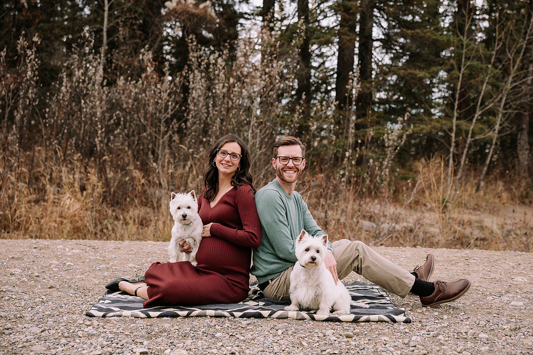 couple sitting on rocks while dressed in fall attire. The woman is pregnant and they are posing for a photoshoot with their dogs.