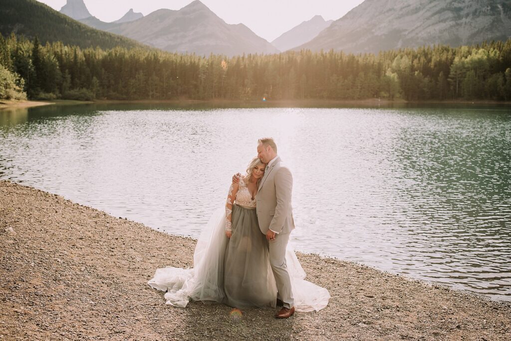 bride and groom snuggling romantically in front of a pond. The lighting is soft and there is a mountain range behind them