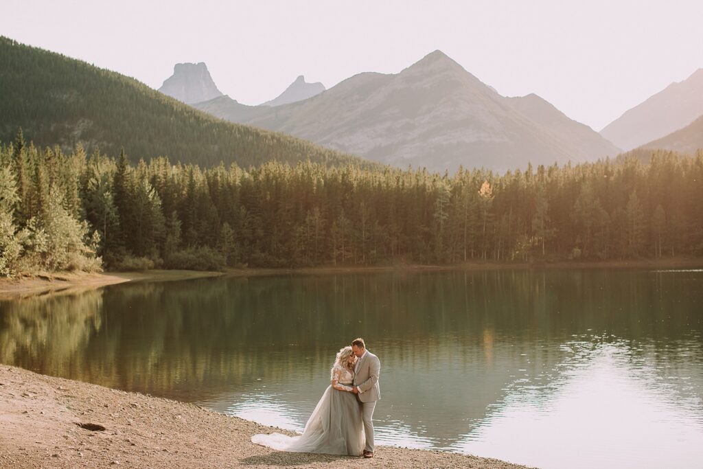 bride and groom snuggling romantically in front of a pond. The lighting is soft and there is a mountain range behind them