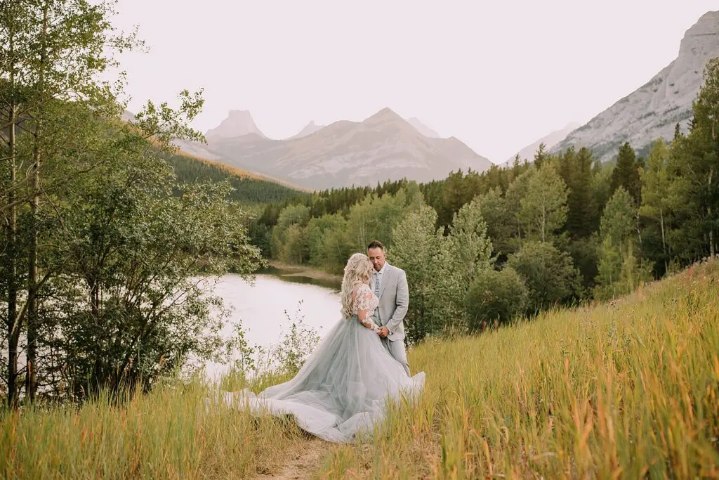 bride and groom snuggling romantically in front of a pond. The lighting is soft and there is a mountain range behind them