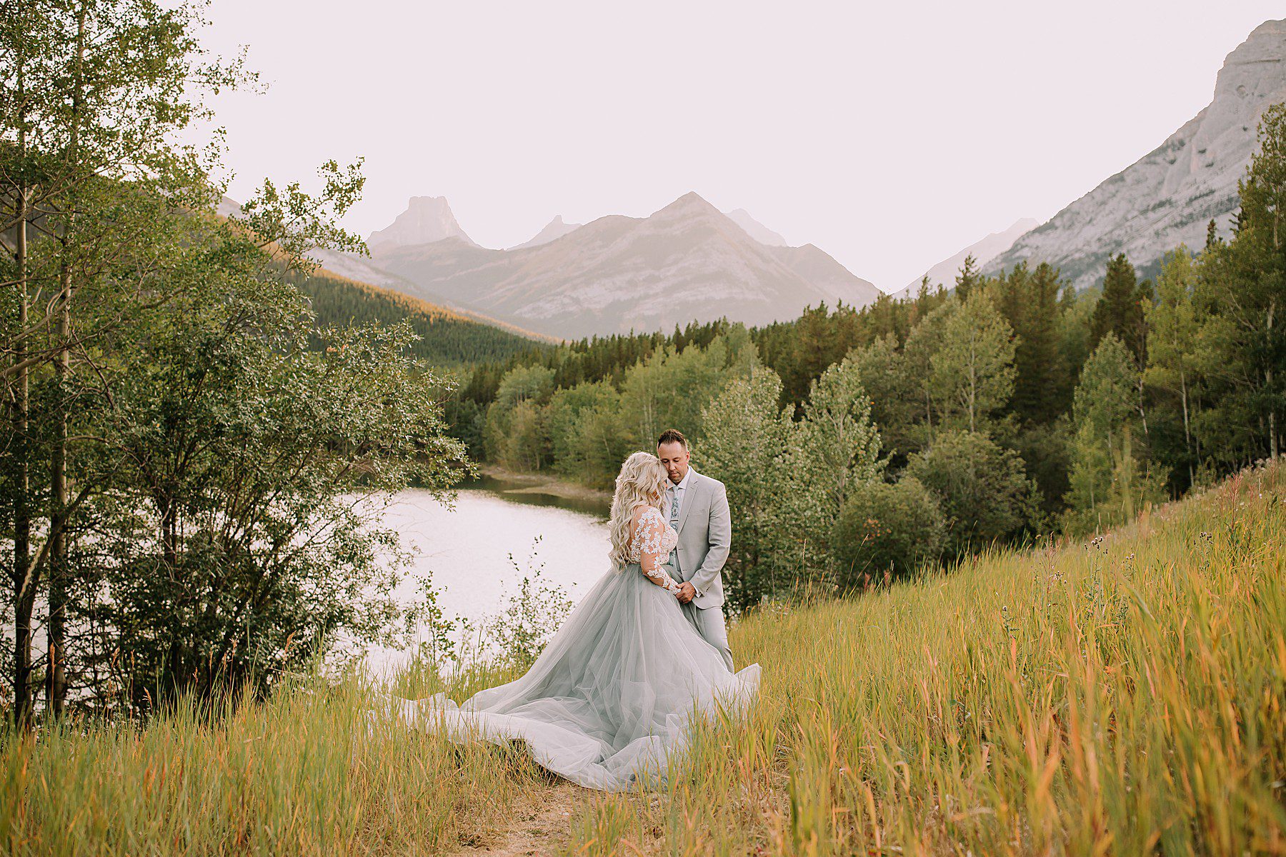 bride and groom snuggling romantically in front of a pond. The lighting is soft and there is a mountain range behind them